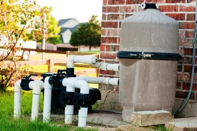 Pool filter system with pipes against a brick wall and green lawn.