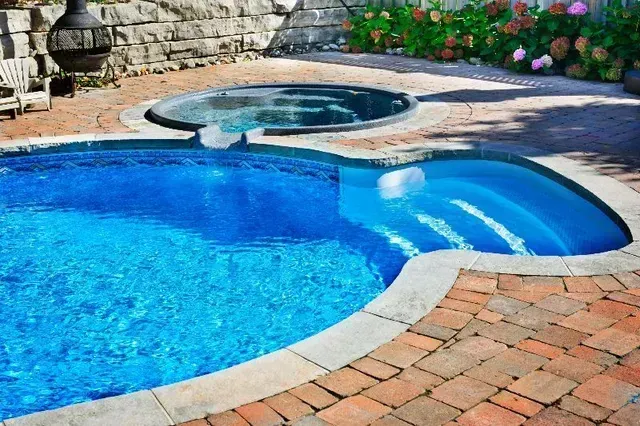 Blue swimming pool and hot tub surrounded by brick patio.
