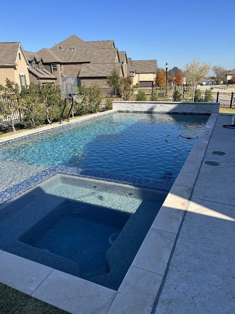 Swimming pool with a connected hot tub, surrounded by stone and lush greenery, houses in the background.