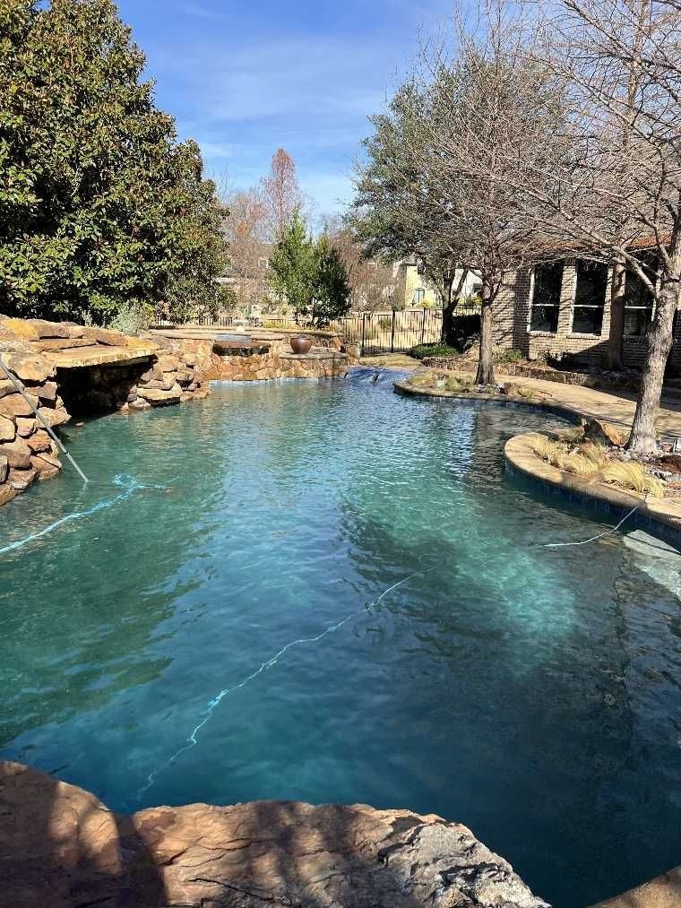 Pool with clear, blue water. A stone waterfall feature is visible. A house is in the background. Sunny day.