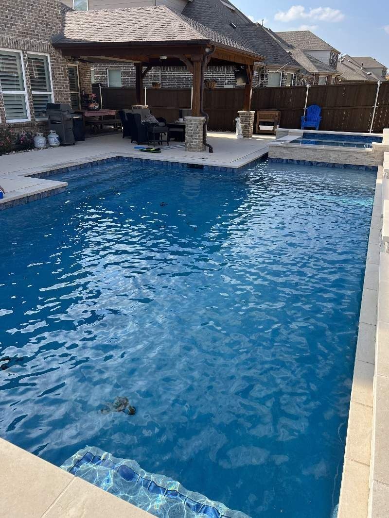 Pool with pergola, stone patio, and spa area in a backyard setting. Blue water, sunny day.