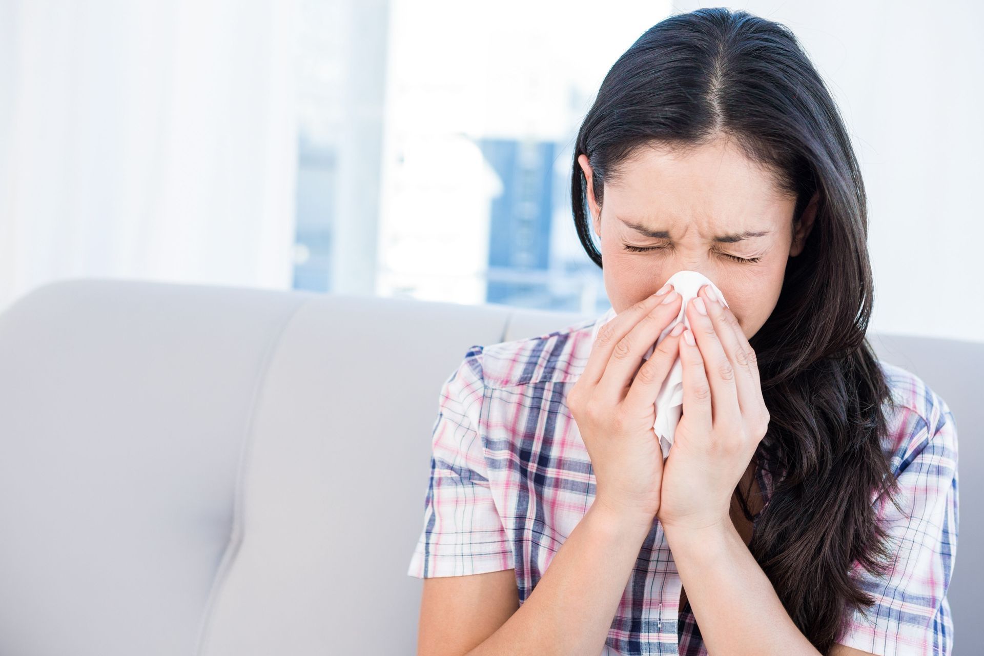Woman on couch blowing her nose with a tissue, eyes closed, indoors.