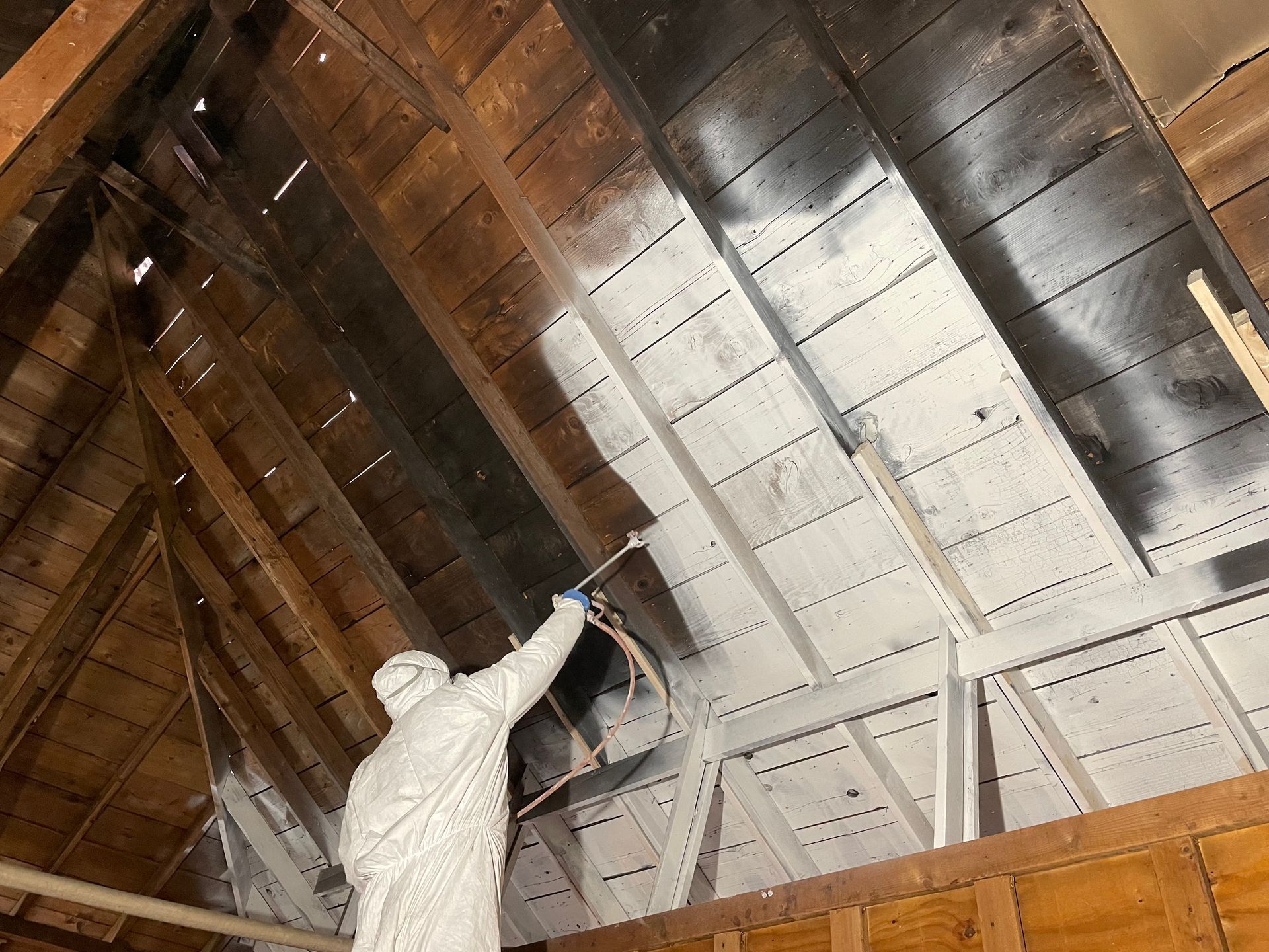 Person in protective suit sprays white coating on wooden attic ceiling.