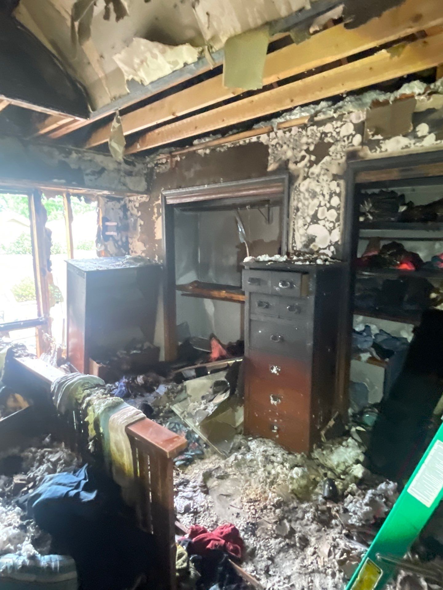 Fire-damaged room: charred furniture, debris, and exposed framing. Walls and ceiling extensively burned, visible window.