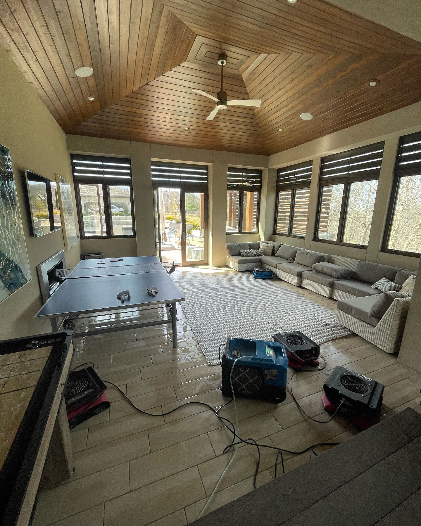 Room with water damage. Dehumidifiers, large sectional, ping pong table, light wood ceiling and floor.