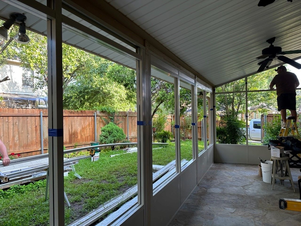 A man is working on a screened in porch with a ceiling fan.