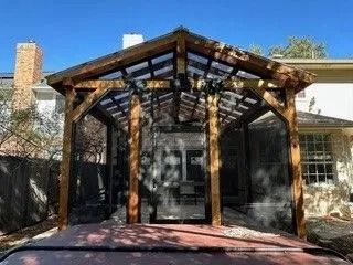 A car is parked under a wooden gazebo in front of a house.