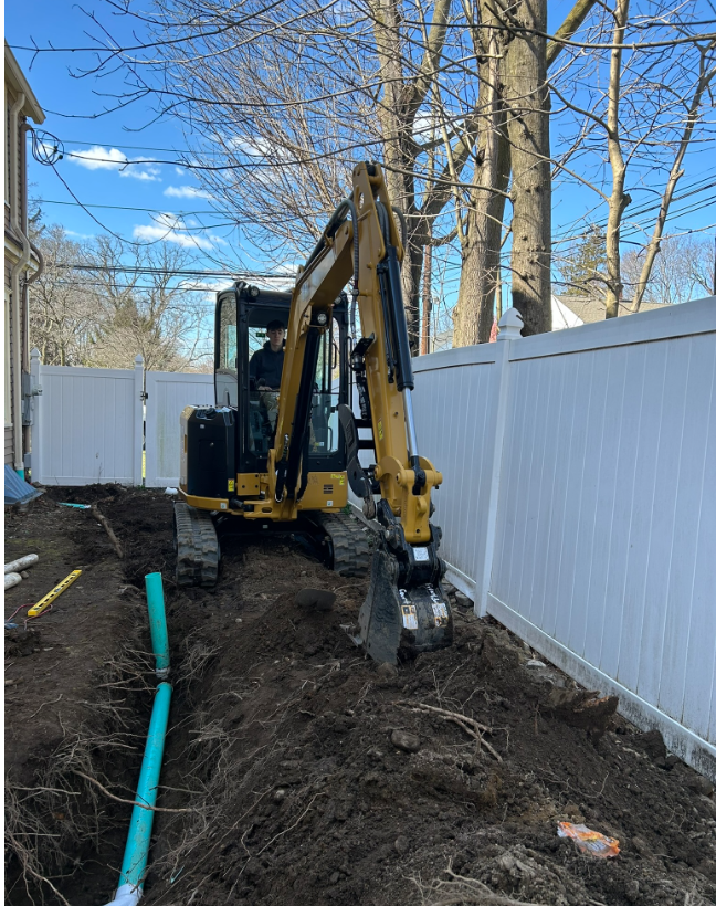 A yellow excavator is digging a hole in the dirt in front of a white fence.