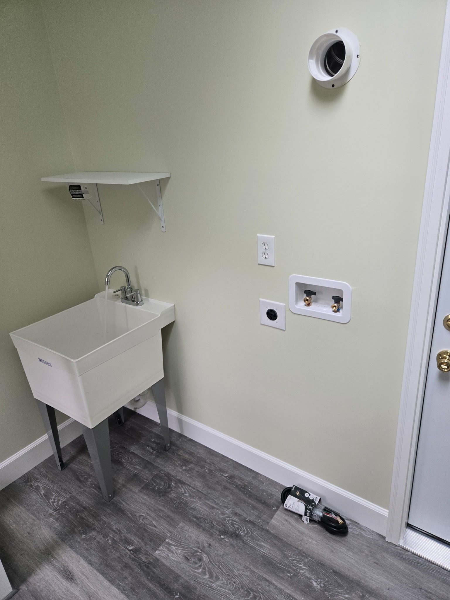 A laundry room wall with a white utility sink, shelf, water hookups, electrical outlets, and a dryer vent port.