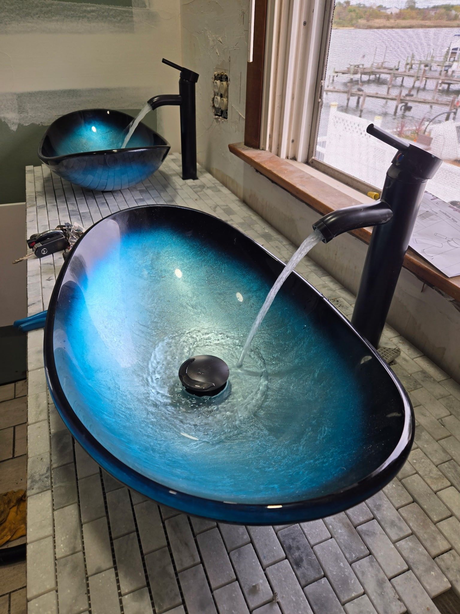Two oval, blue-to-black gradient glass vessel sinks with black faucets running water, set on a light-colored tiled counter.
