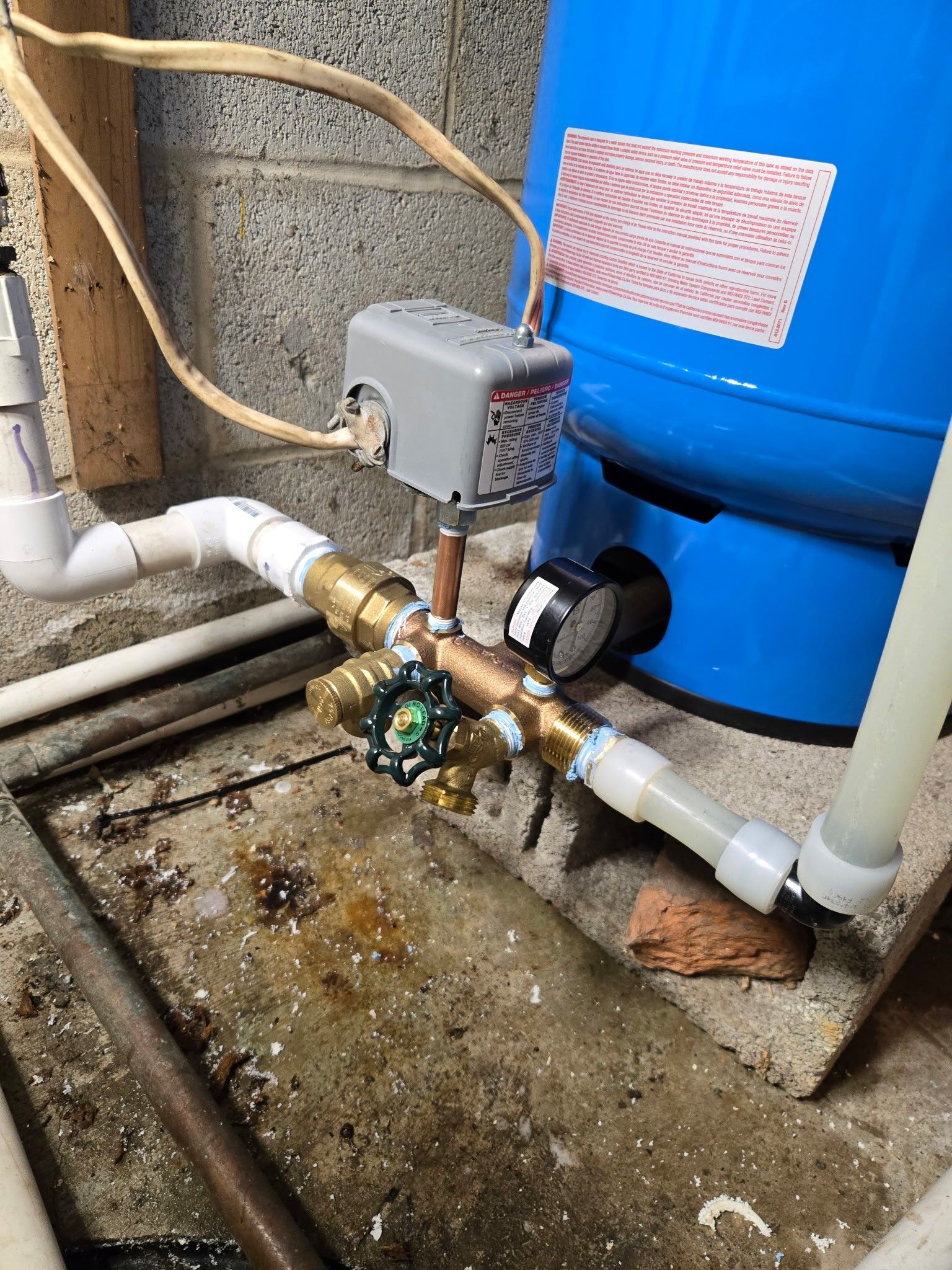 A blue well pressure tank connected to white PVC pipes, a brass manifold, a gray pressure switch, and a round gauge.