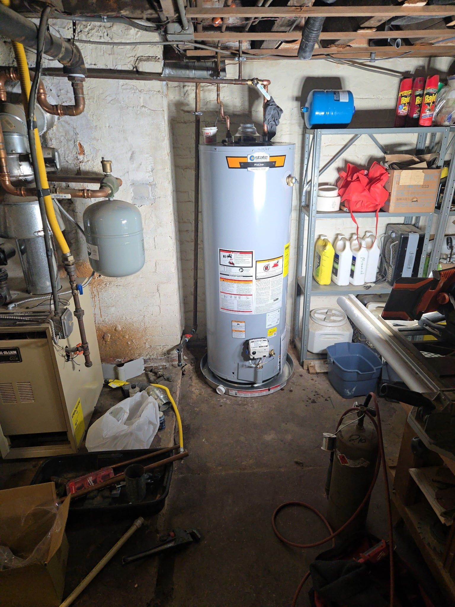 A gray water heater stands in a cluttered basement next to a storage shelf and various home maintenance equipment.