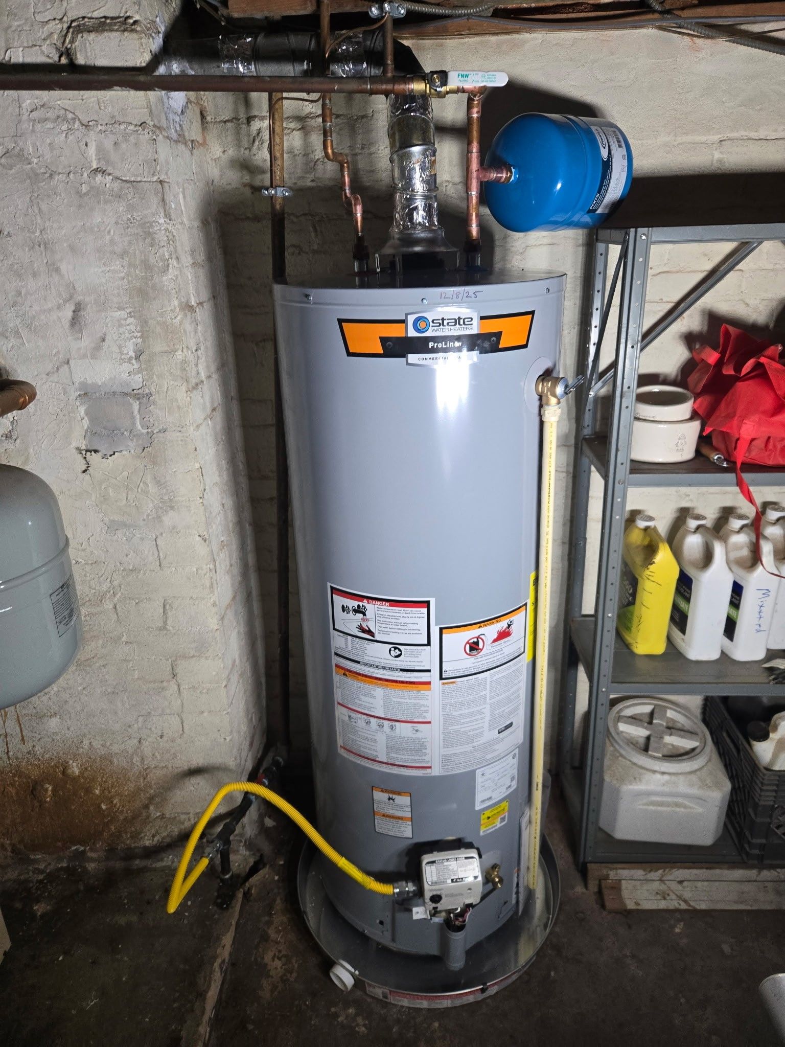A grey gas water heater in a basement, connected to overhead copper pipes and a blue expansion tank on a metal shelf.