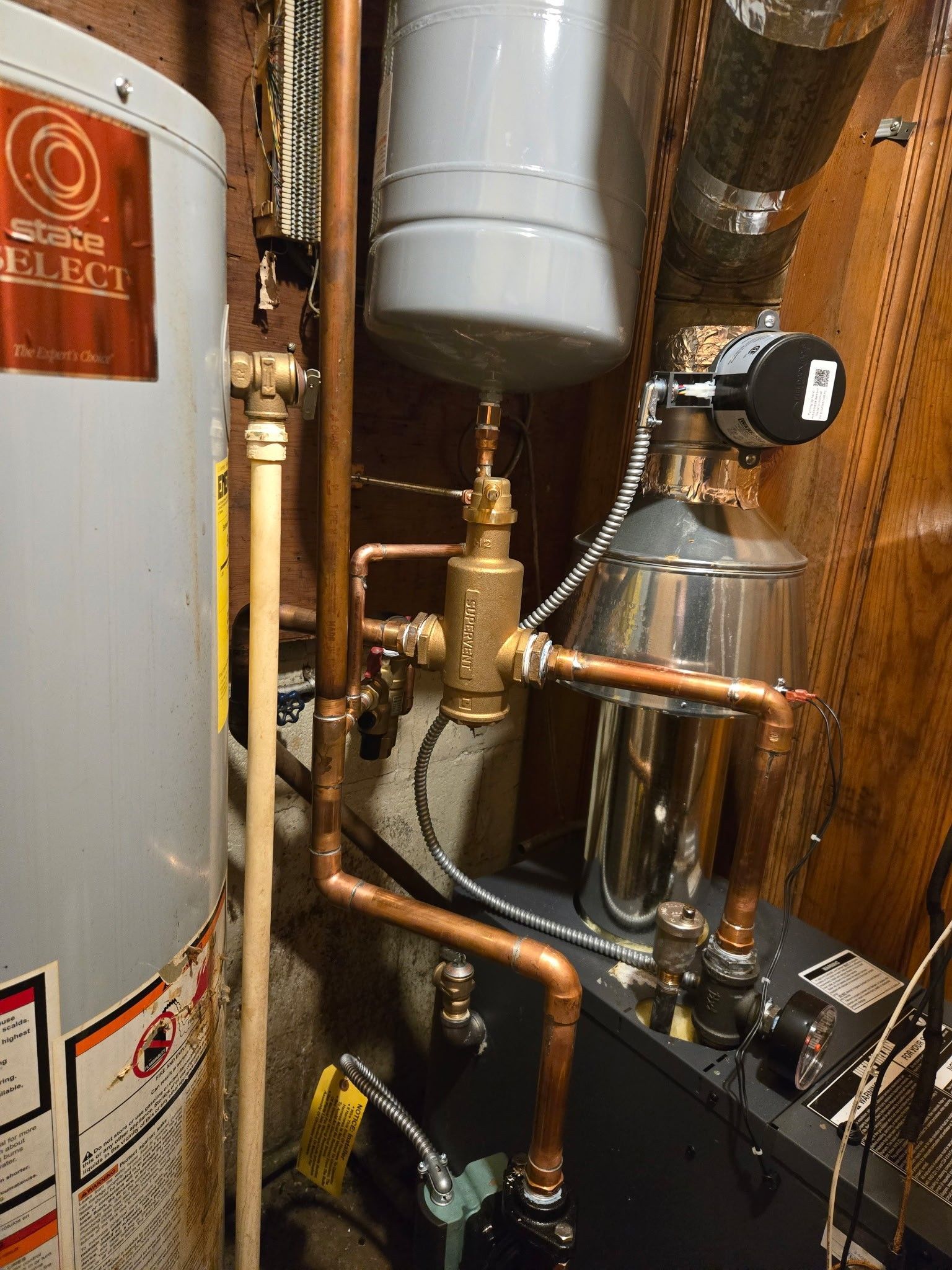 A water heater and boiler setup with copper piping, an expansion tank, and a pressure-reducing valve in a utility area.