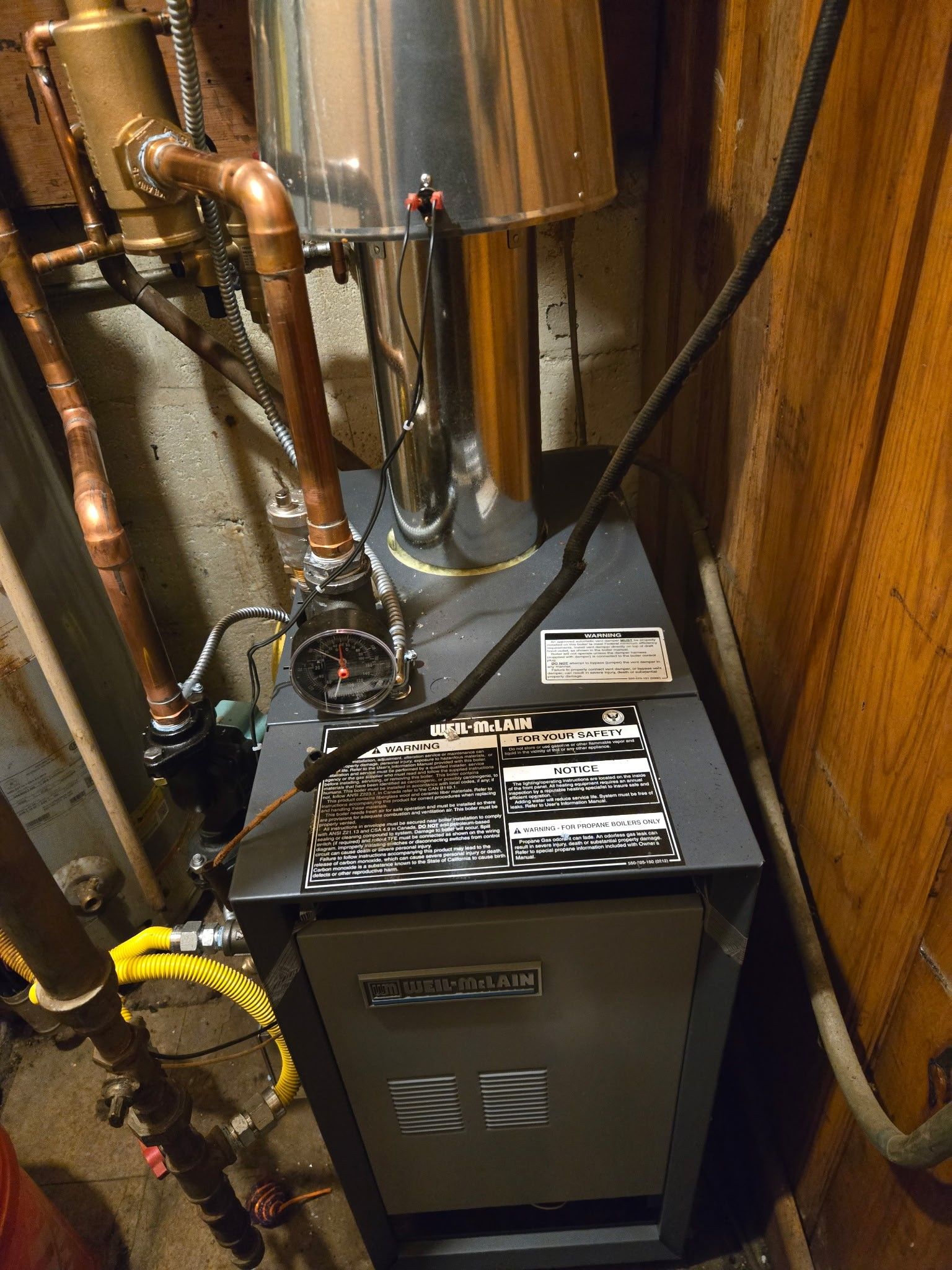 A metallic residential boiler with copper piping and a chrome vent pipe against a wood-paneled wall.