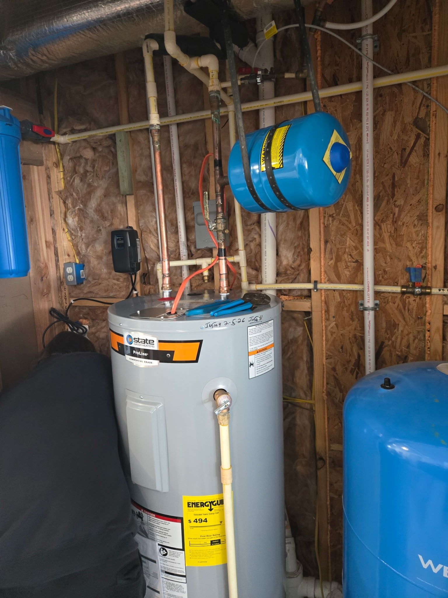 A water heater with a blue expansion tank mounted on a wooden wall next to a large blue water storage tank.
