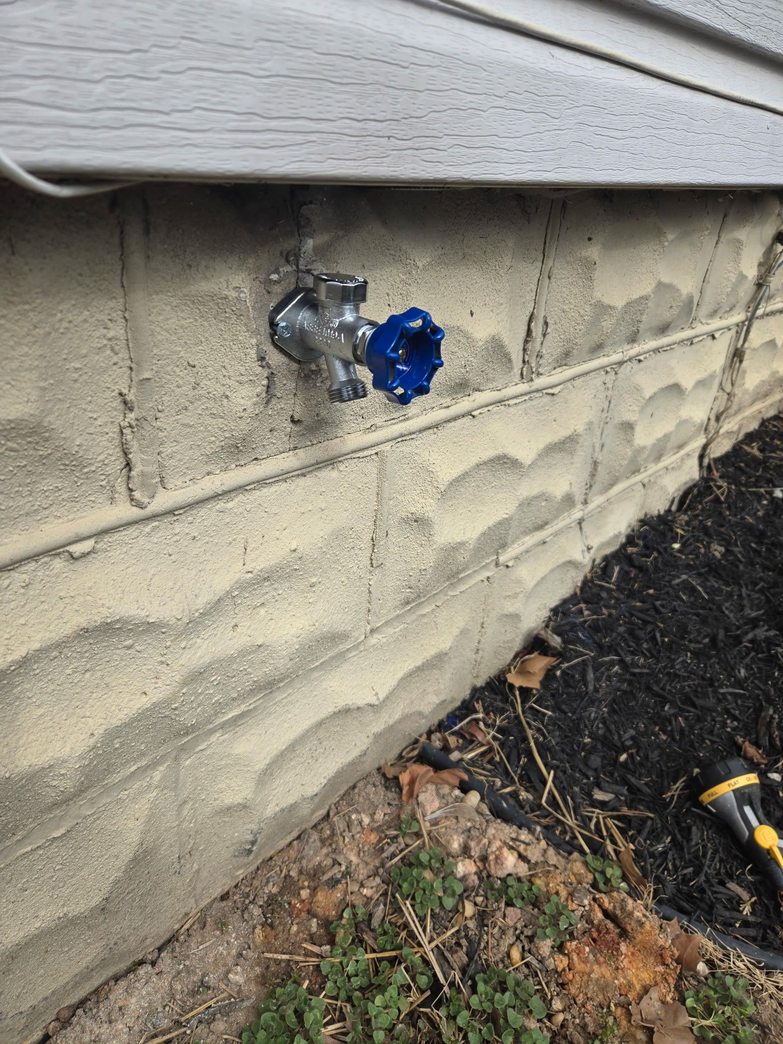 A metal outdoor water faucet with a blue handle attached to a light-colored concrete foundation wall.