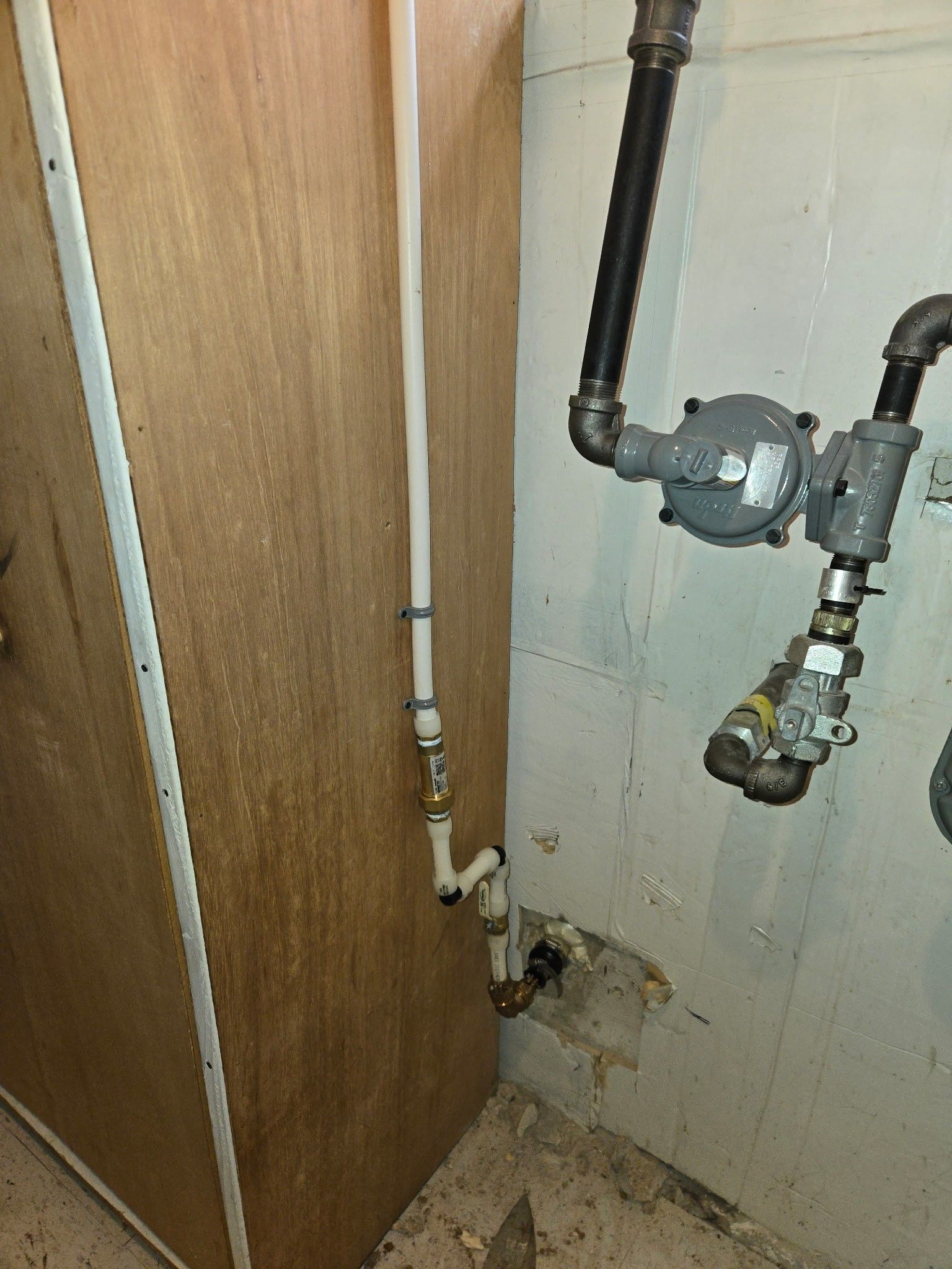 White PEX piping running along a wooden cabinet next to a black and gray natural gas regulator assembly on a concrete wall.