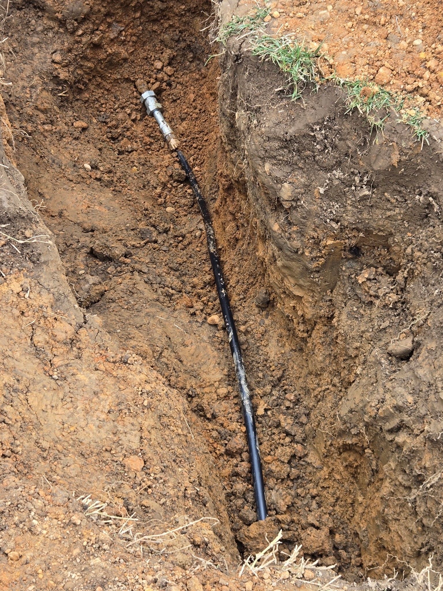 A black pipe with a metal coupling lies inside a narrow, freshly dug dirt trench.