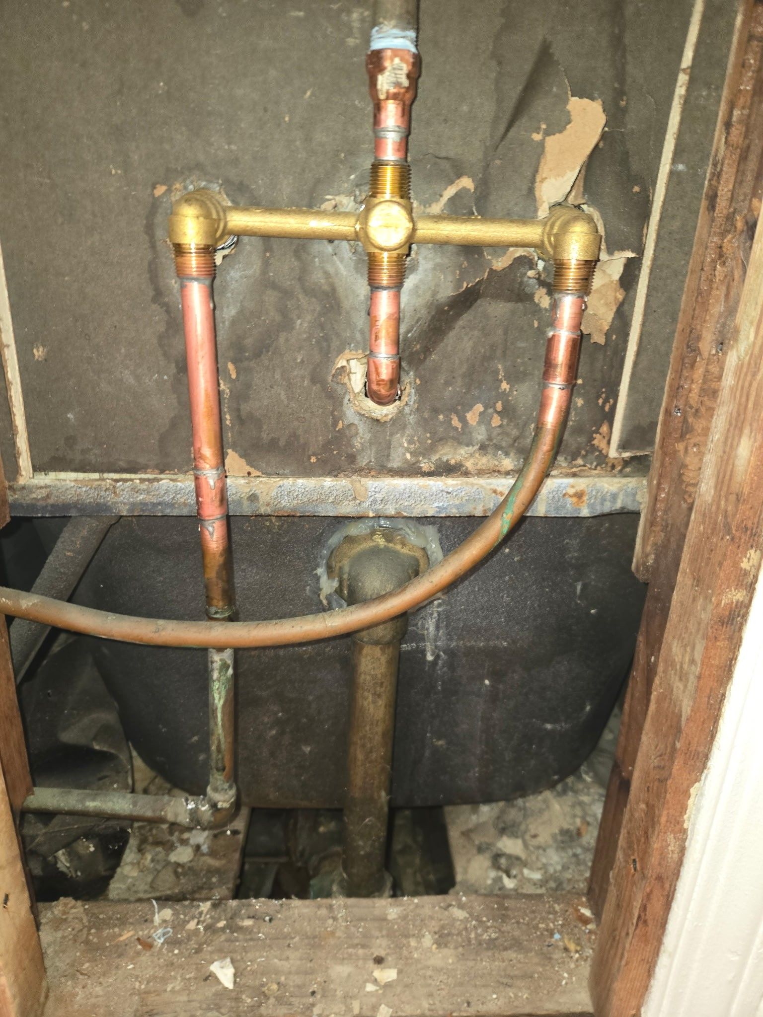 Copper plumbing pipes connect to a brass valve assembly behind a wall, positioned above a black sink or tub basin.