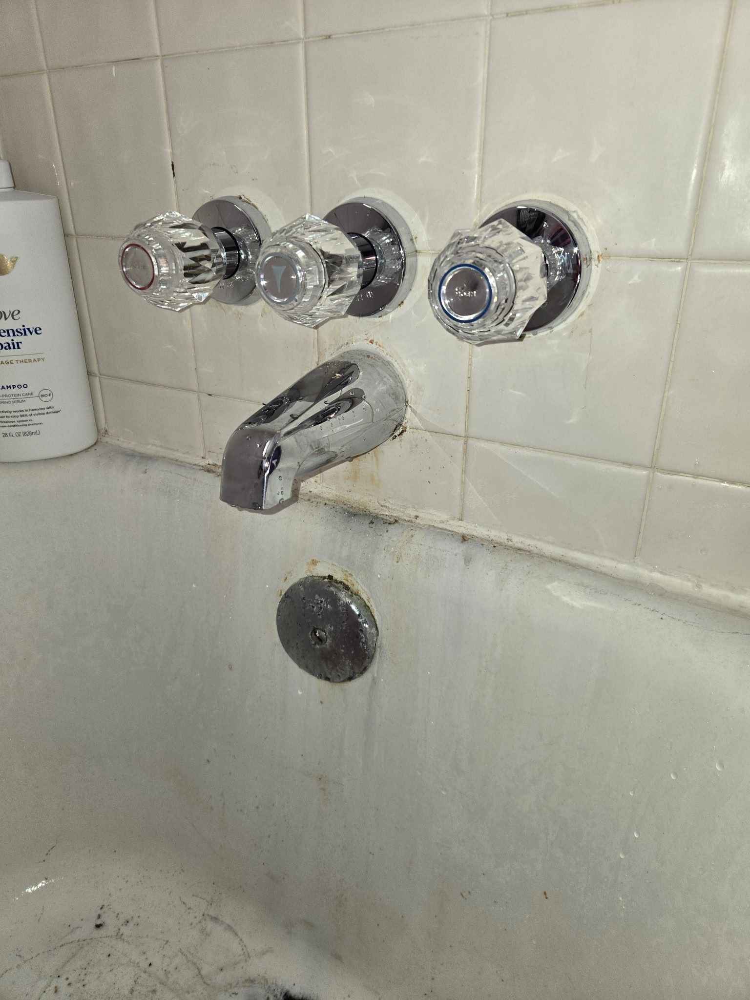 Bathtub faucet with three clear acrylic handles mounted on a tiled wall above a stained tub.