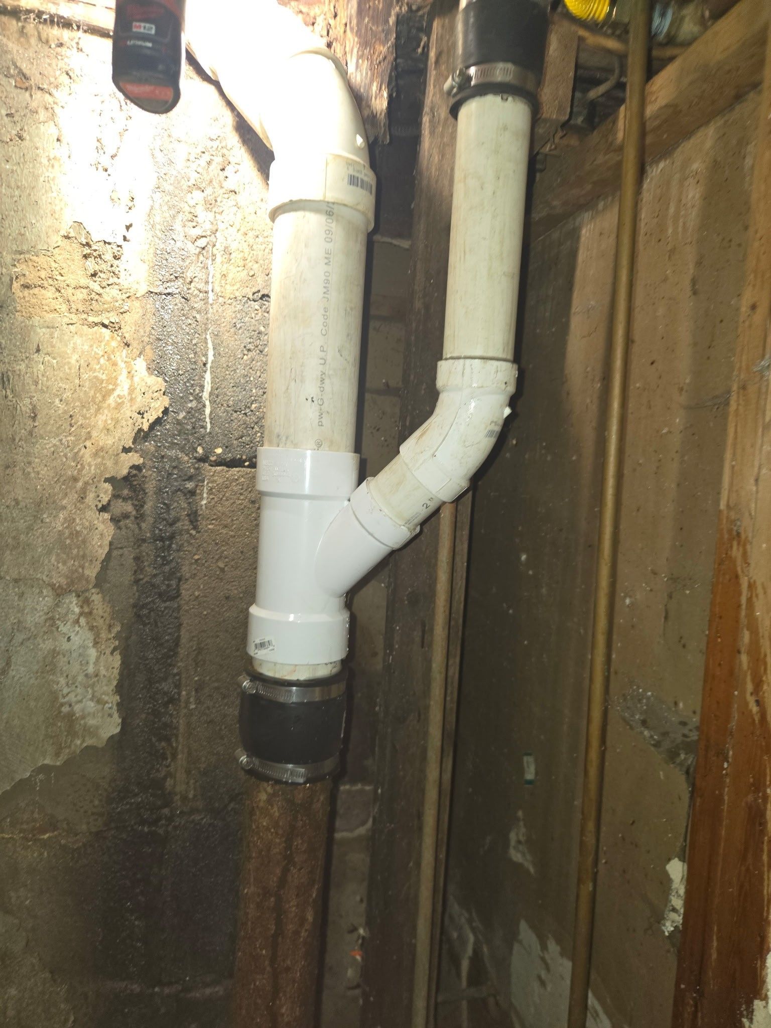 A white PVC plumbing Y-fitting connecting two pipes against a textured, dark wall, joined by black rubber couplings.