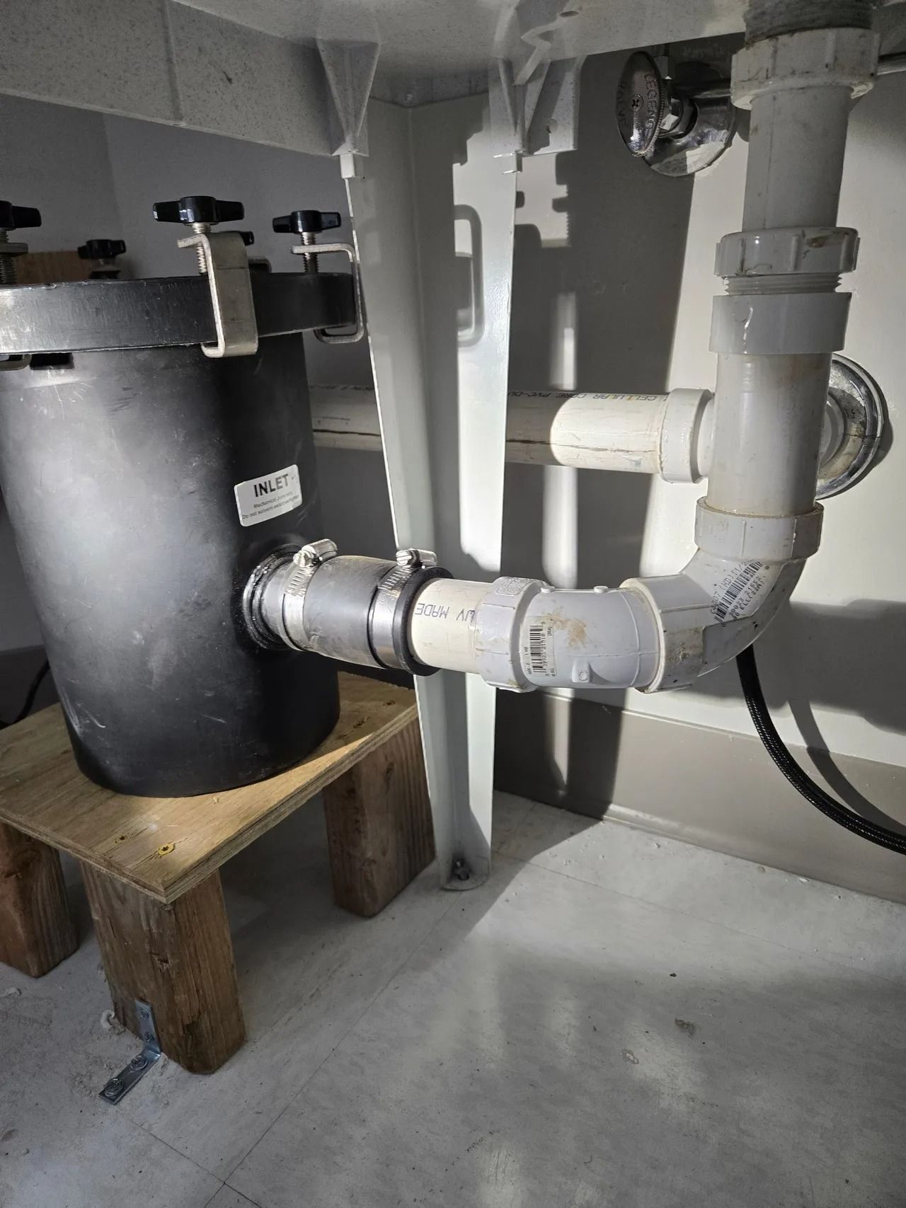 Black water filtration tank with plumbing under a white sink, supported by a wooden stool, against a gray wall.