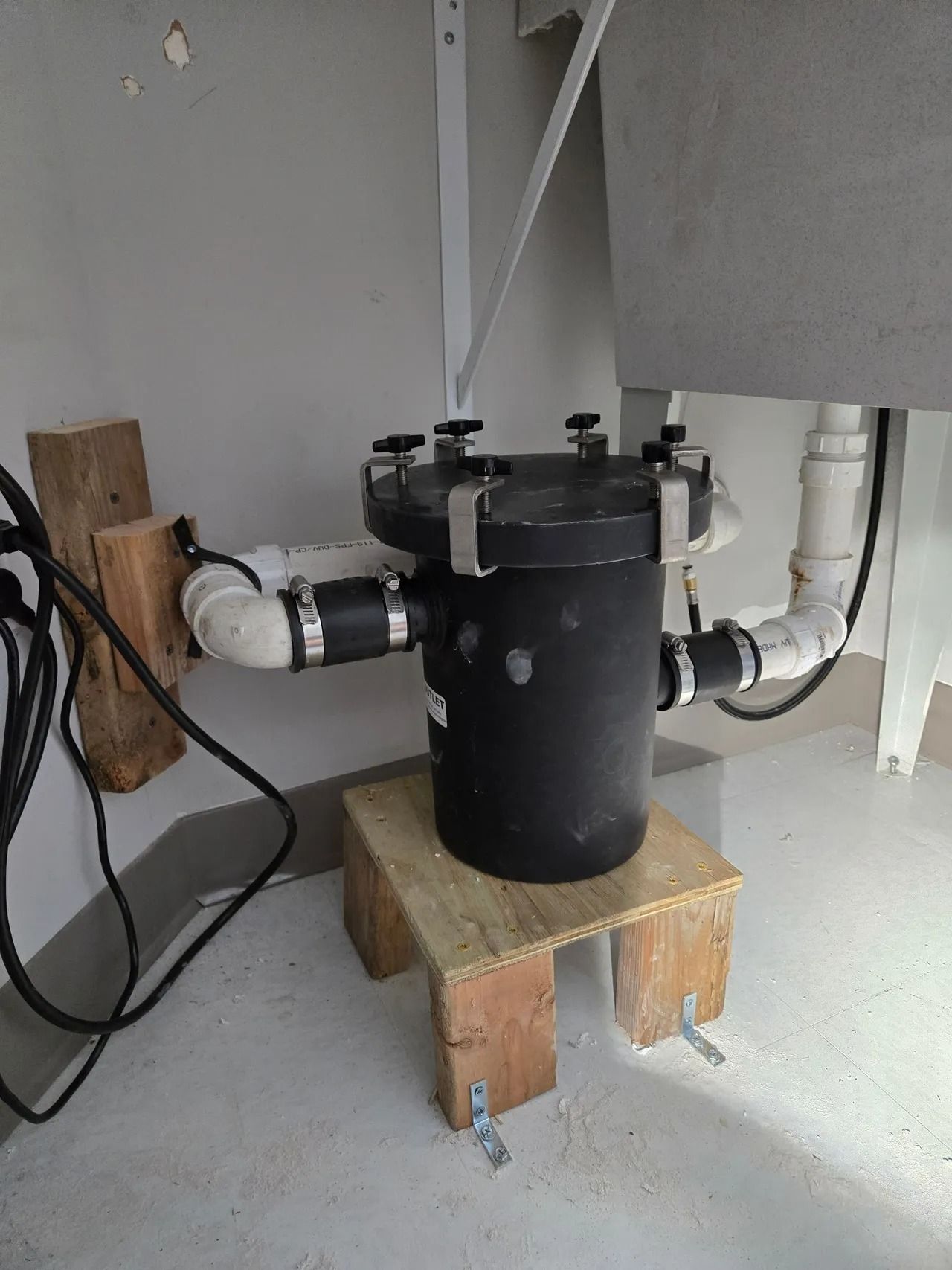 Black filter on a wooden stand, connected by white pipes; in a corner near a sink.
