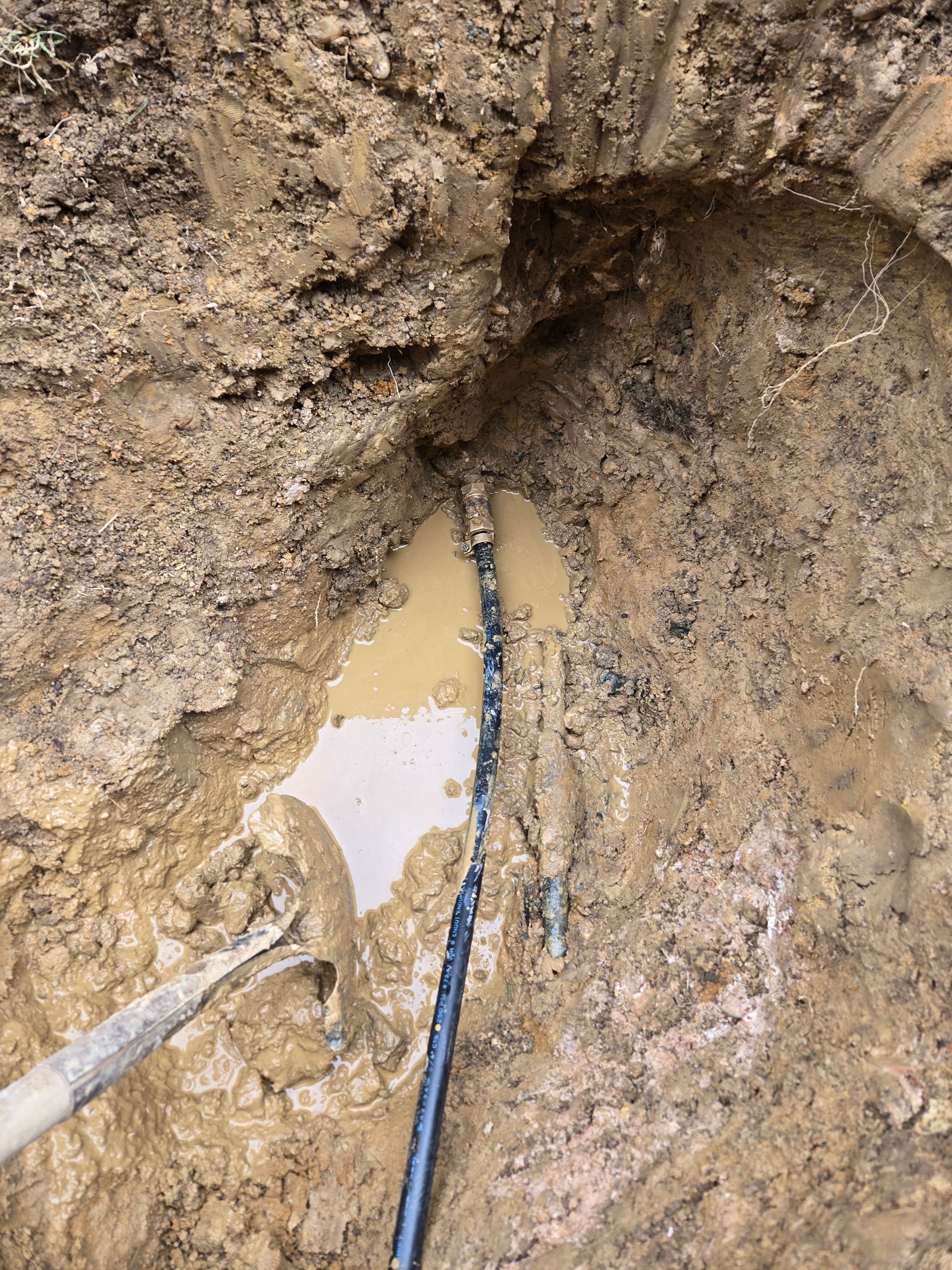 A muddy hole with a pipe and standing water. Shovel rests in the mud.