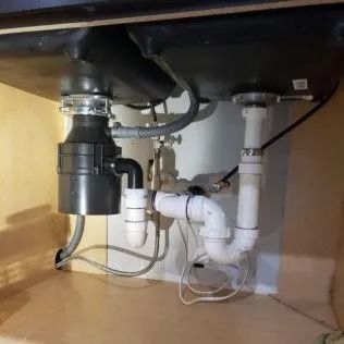Underside of a kitchen sink with a garbage disposal and plumbing visible in a light wood cabinet.