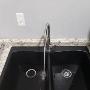 Black double sink with running water from a silver faucet; gray countertop and wall.