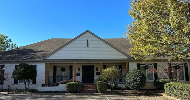 A single-story, white brick house with a triangular roof gable, a central front entrance, and surrounding landscaping.