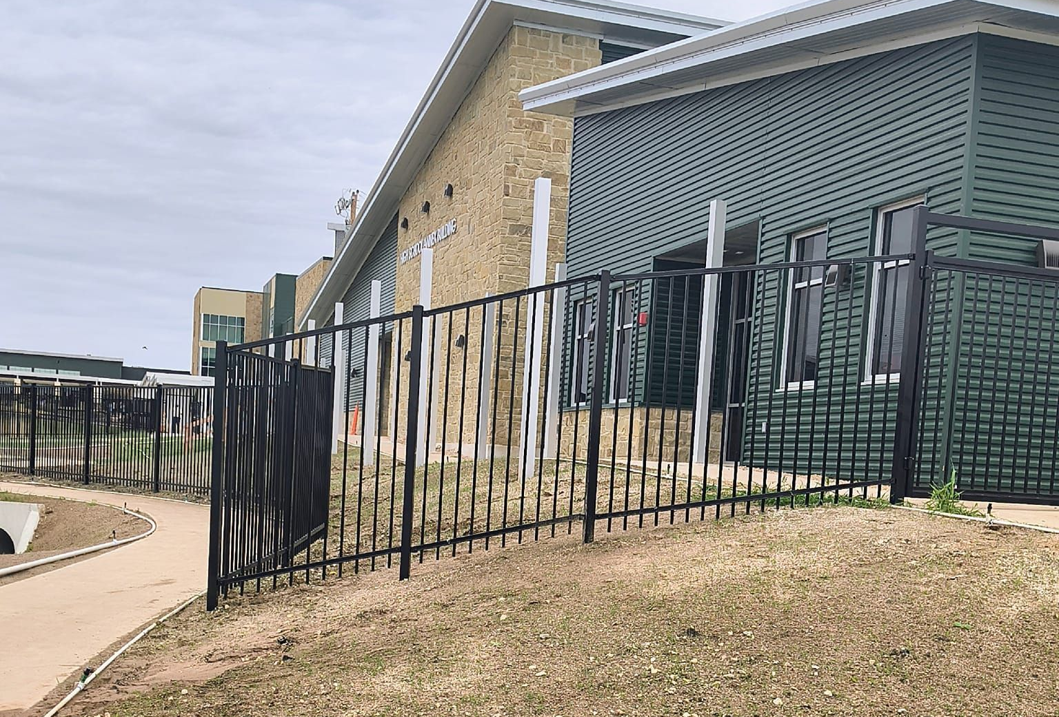 Black metal fence along a green building with windows and a dirt path.