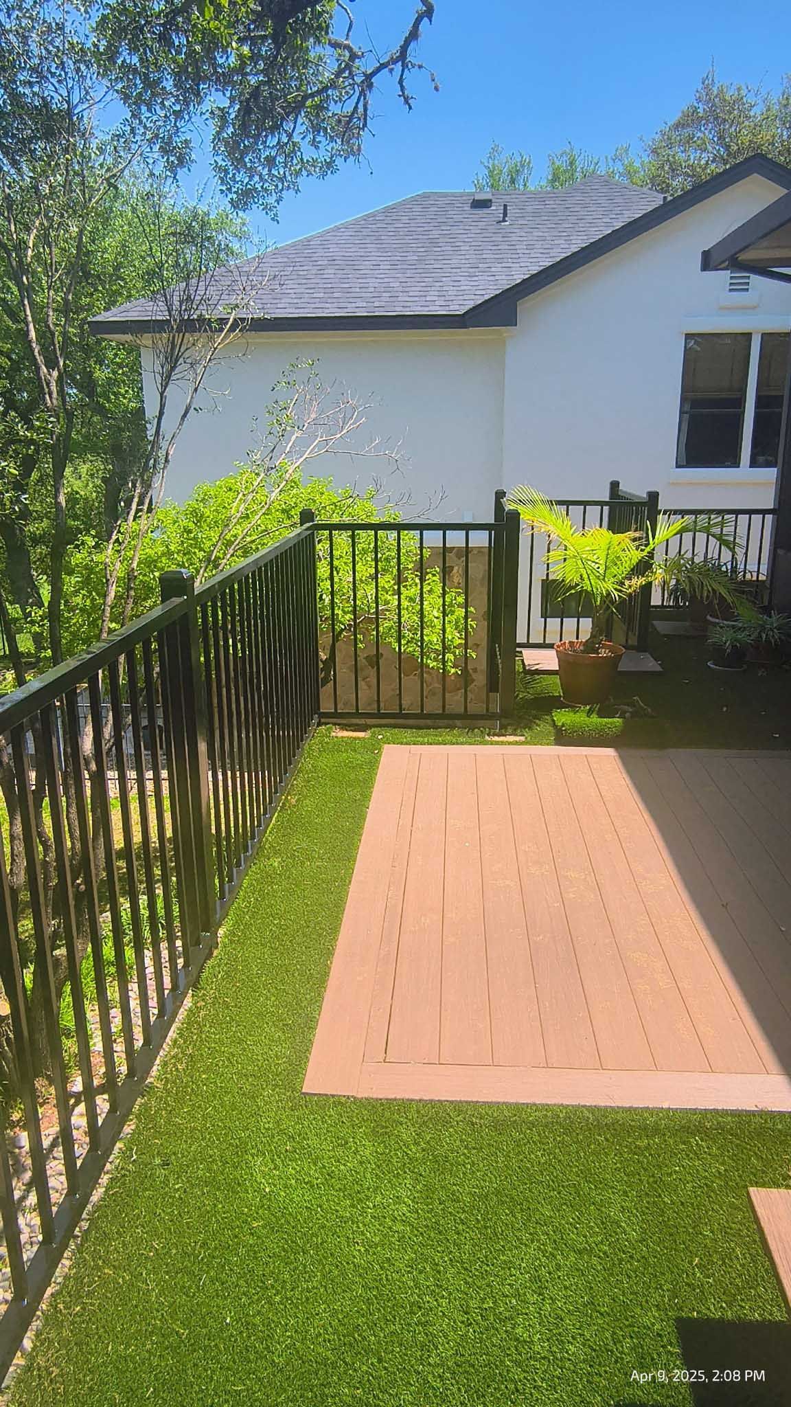 A backyard patio with artificial grass and a black fence, with a white house in the background.