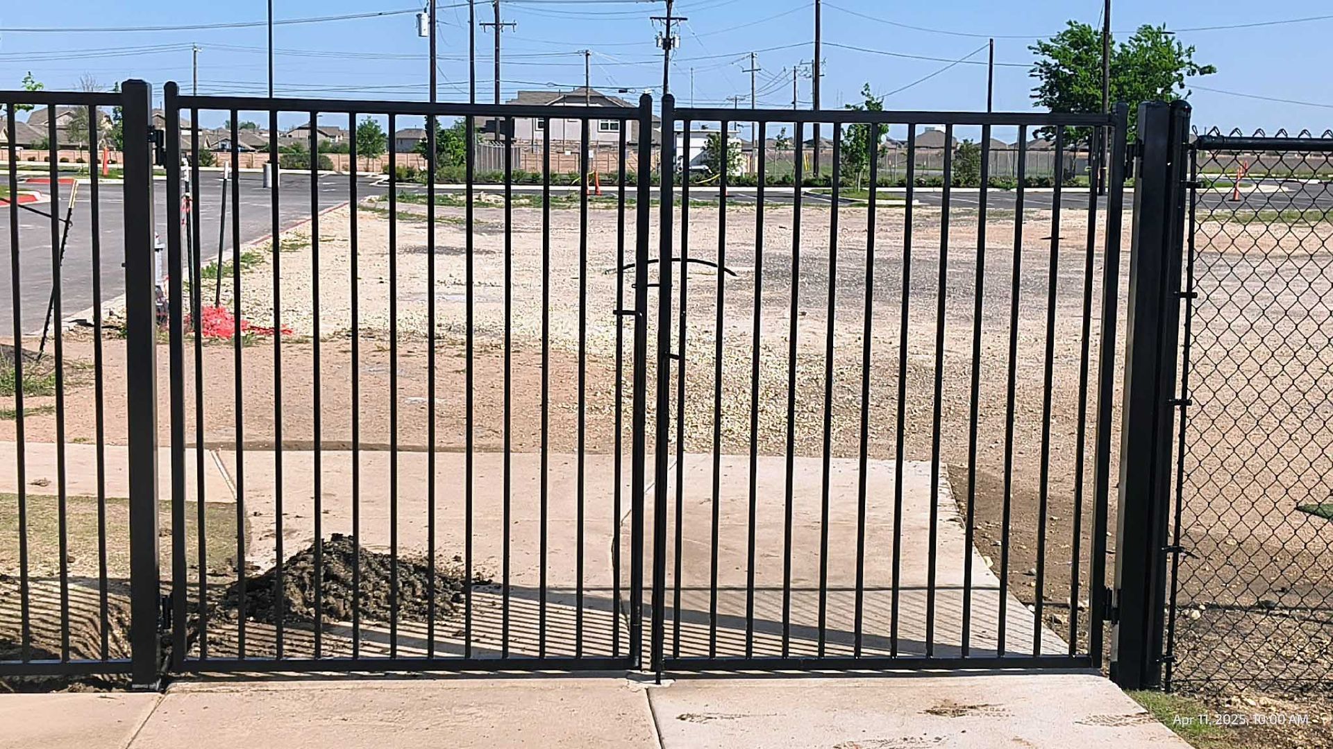 Black metal gates open to a vacant lot, next to a chain-link fence.