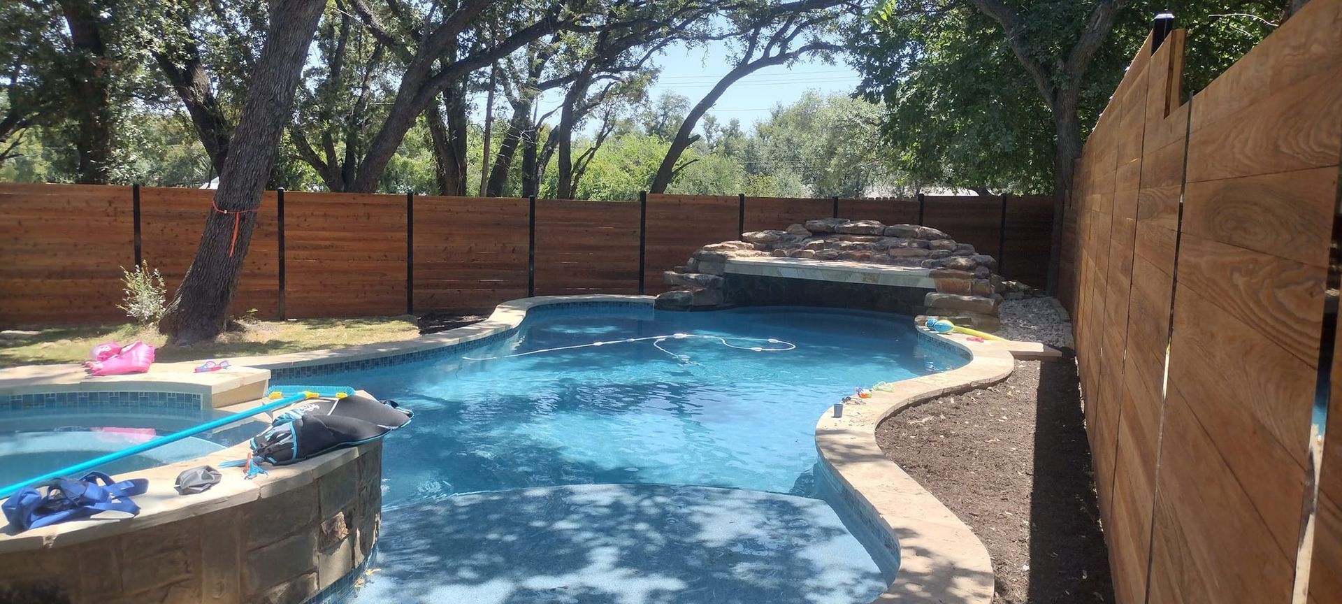 A backyard swimming pool with a waterfall, surrounded by a wooden fence and trees.