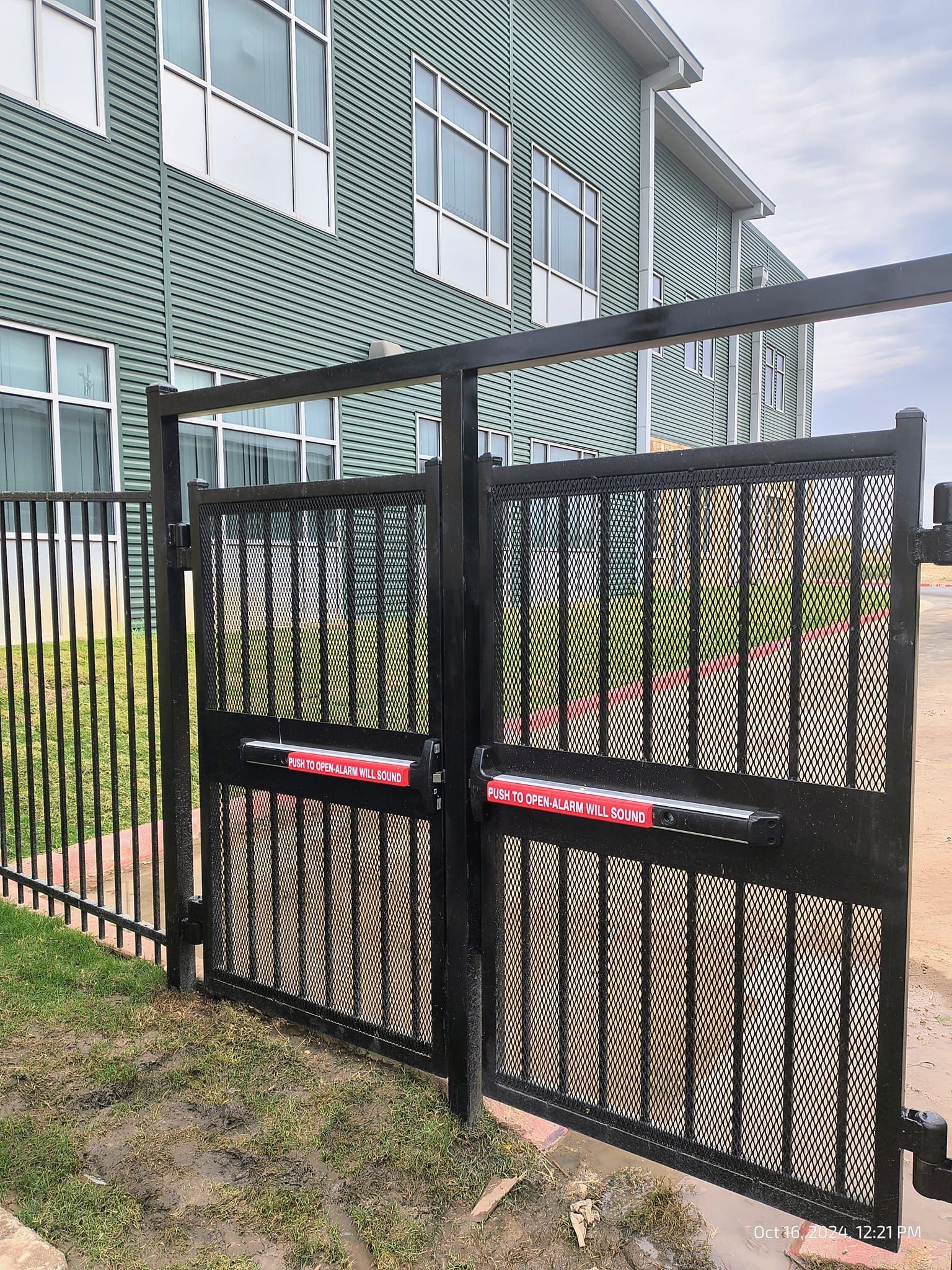 Black metal gate with mesh design and red signs in front of a green building with windows.