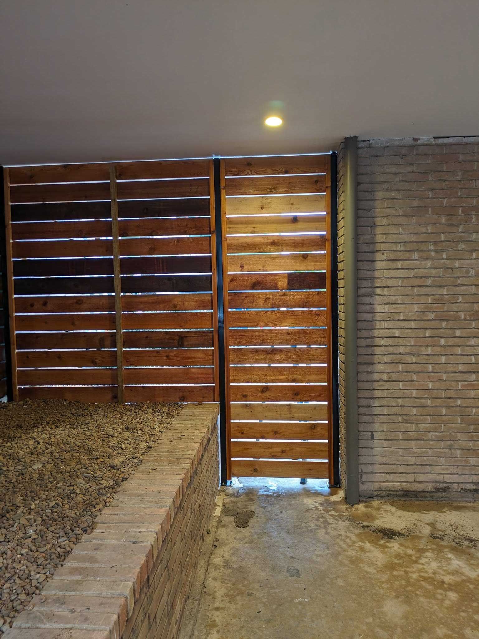 Wooden gate with horizontal slats, partially open, next to a brick wall.