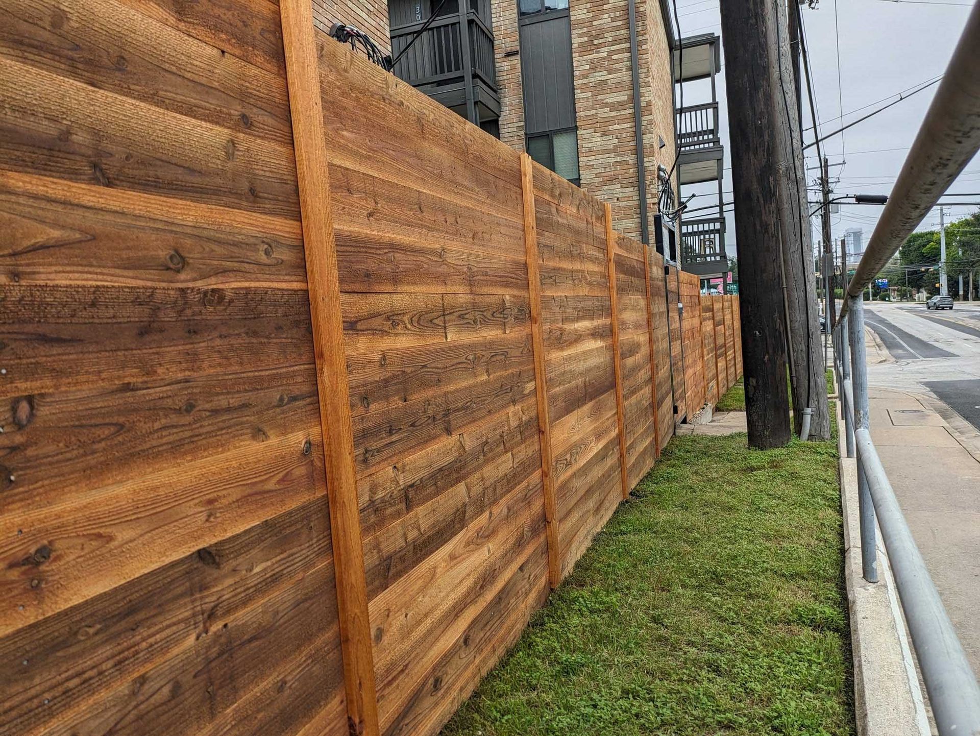 Wooden privacy fence alongside a sidewalk and grassy strip near a building and utility pole.