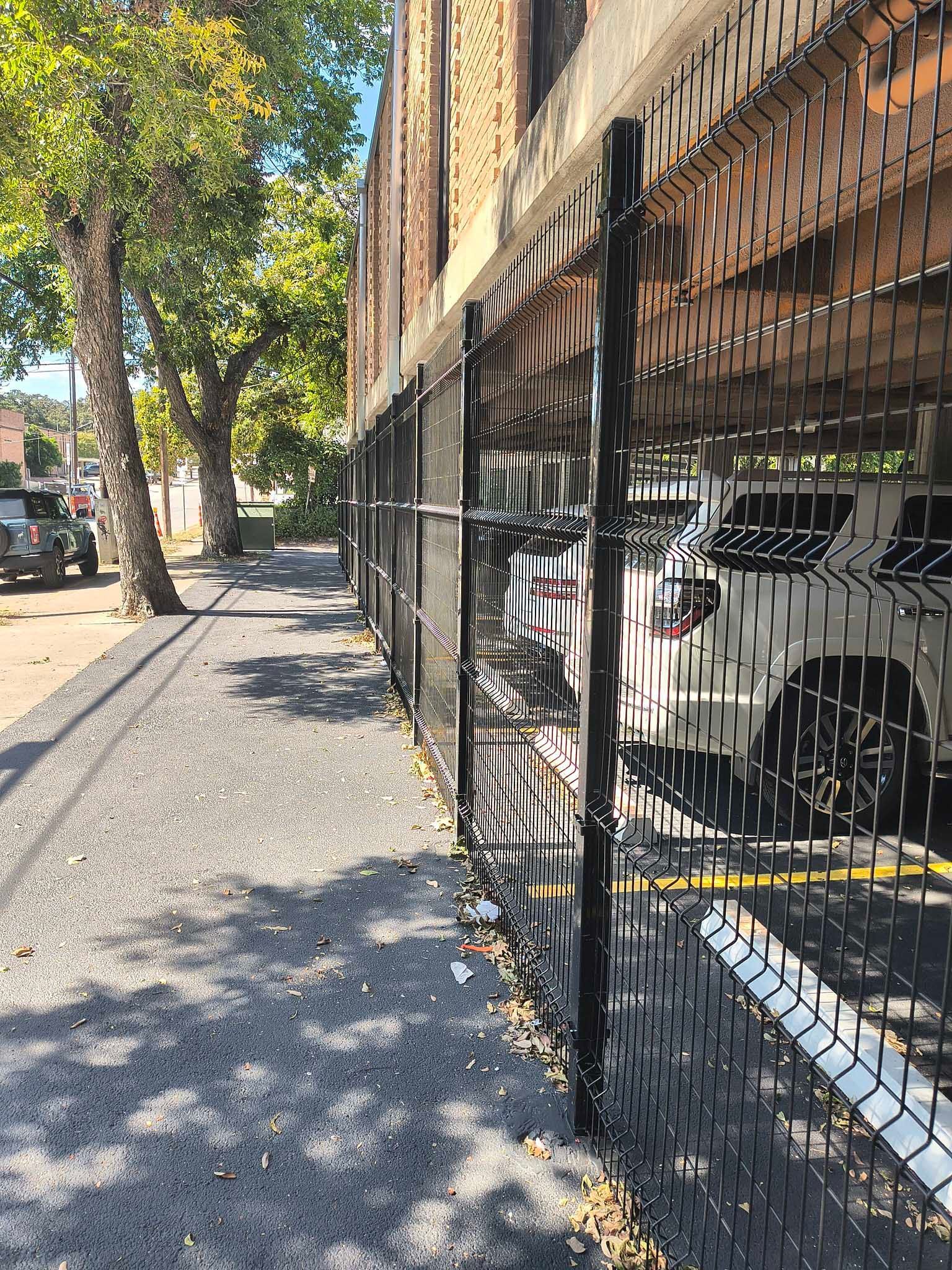 Black fence along a sidewalk next to a building. Cars are visible behind the fence and trees in the background.