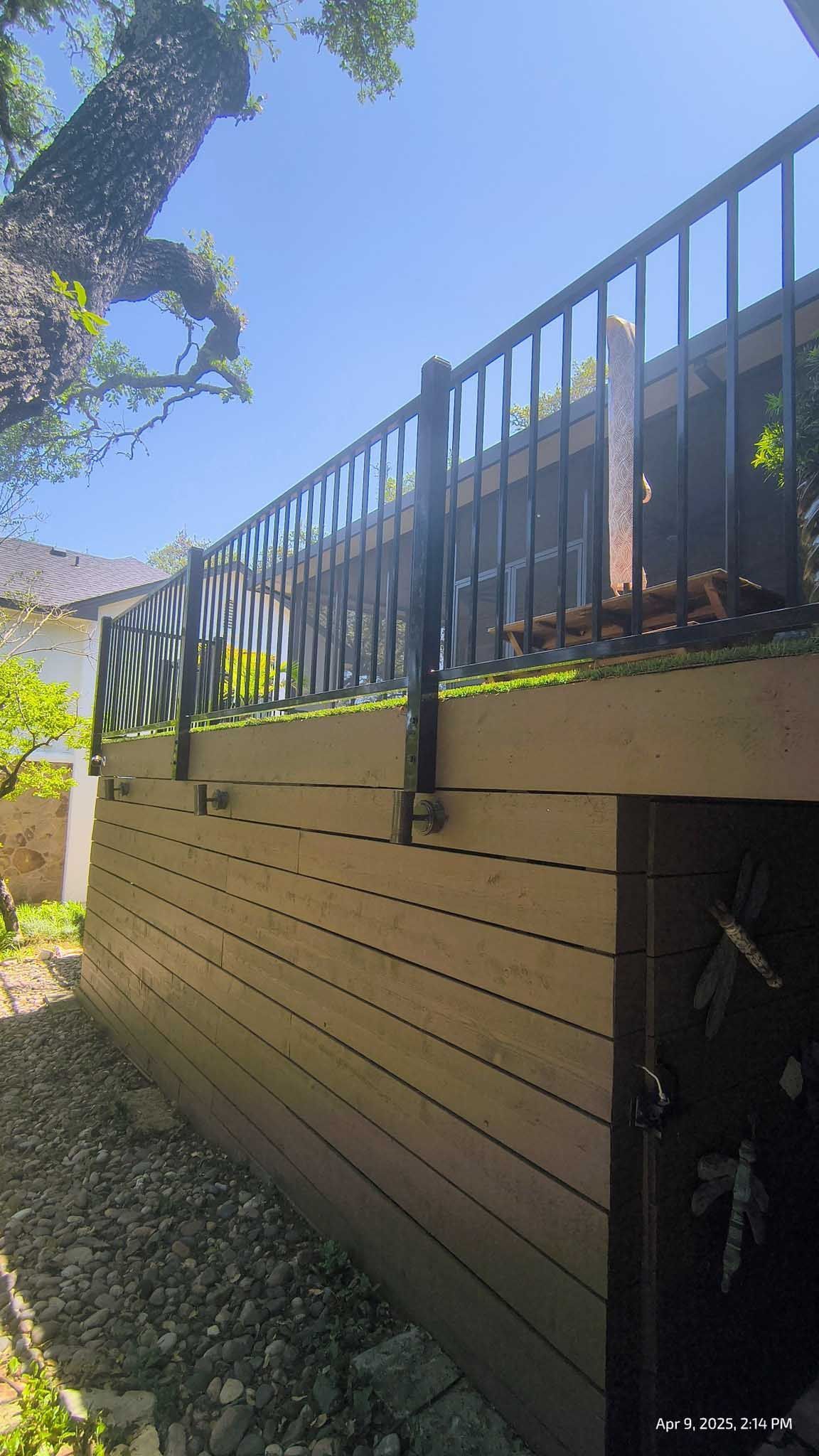 Wooden deck with black railing against a bright blue sky, surrounded by greenery.