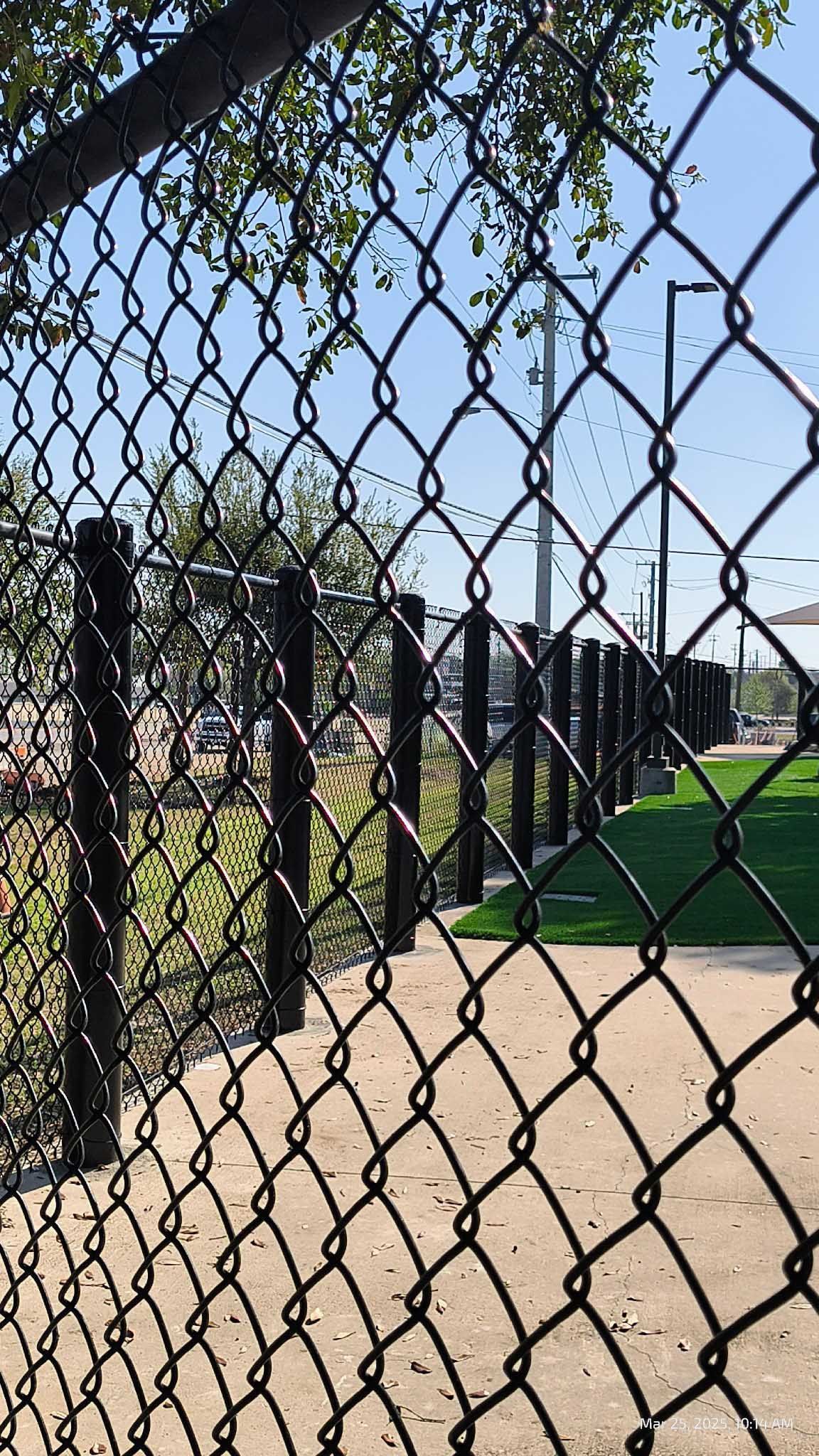 Chain-link fence in foreground, path and park beyond. Blue sky.