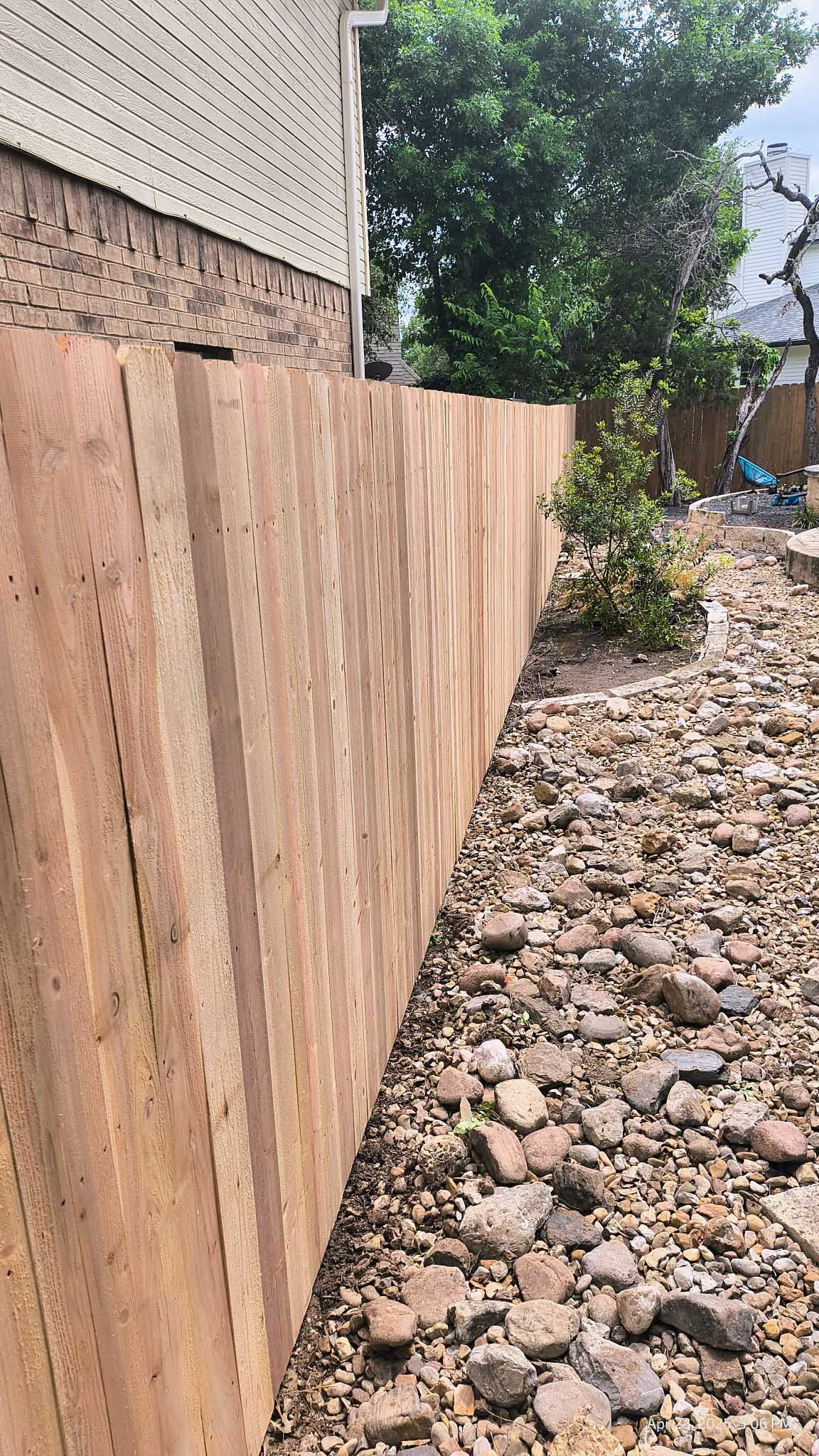 Wooden fence along side a building, next to a rocky patch with some greenery.