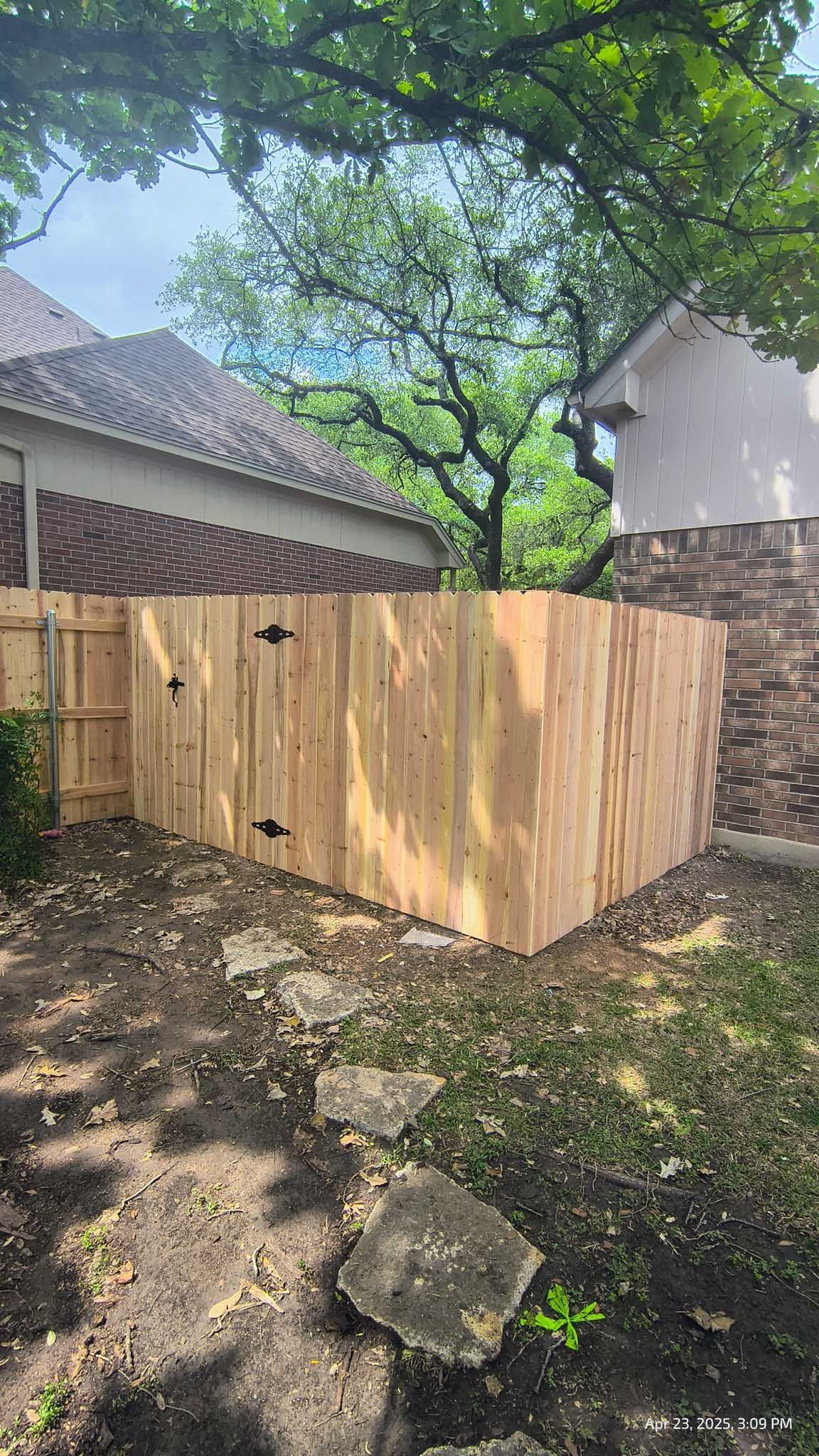 New wooden fence in a yard next to a brick building and a tiled roof, with trees in the background.