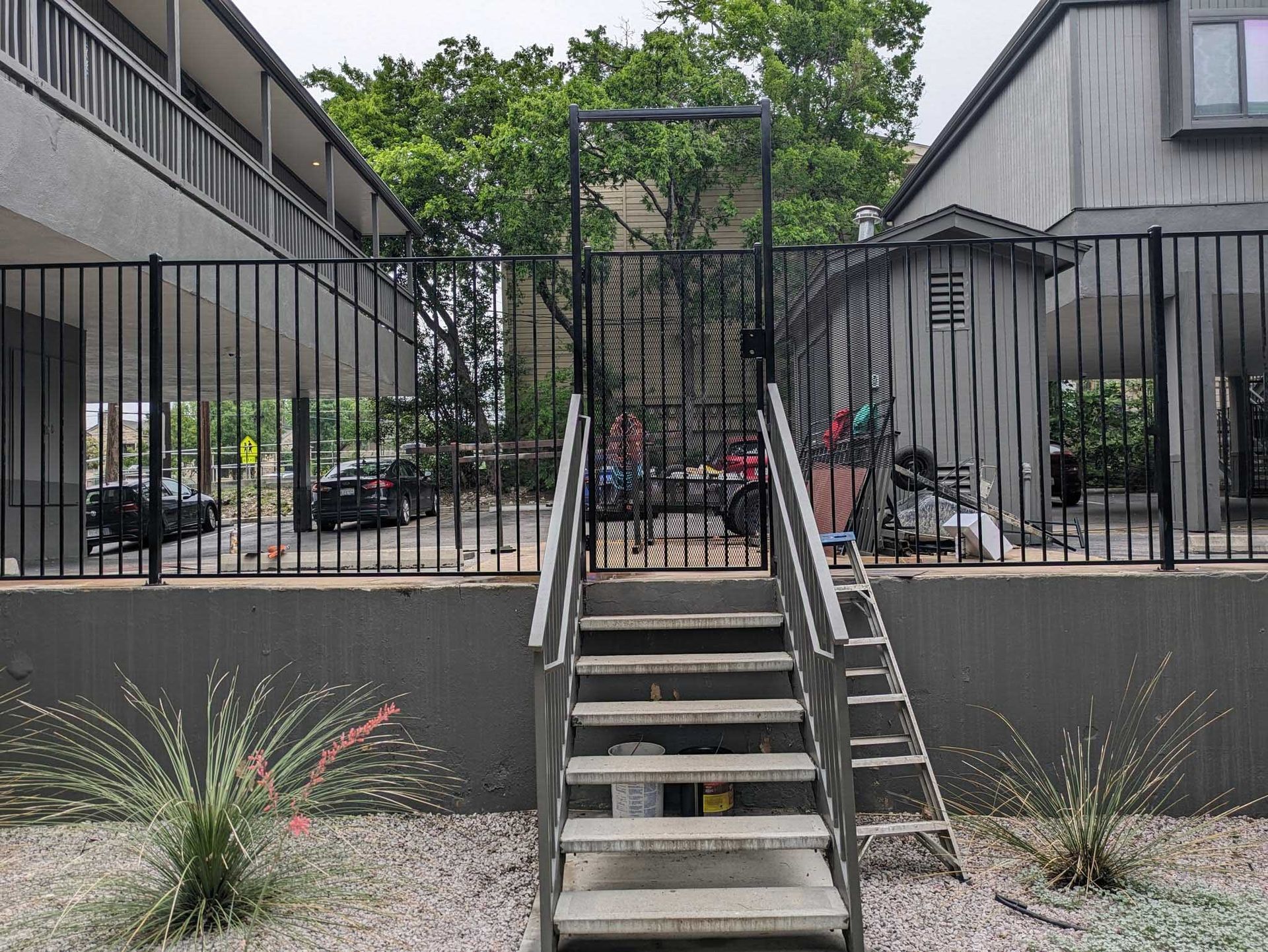 Stairway leading to a black metal gate in a fenced area. A small shed and parking are visible behind.