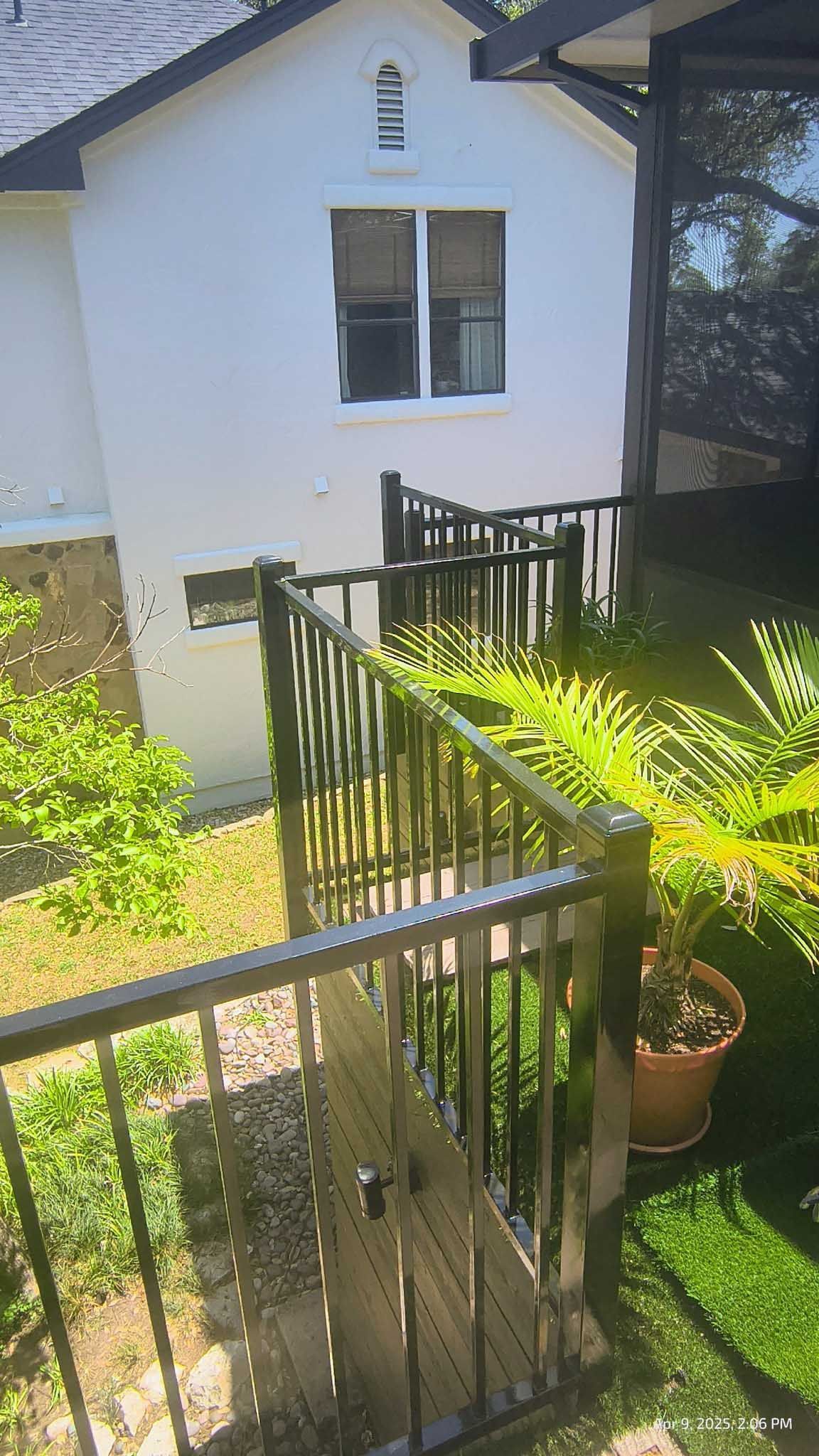 Black metal railing in front of white building with window and door. Plants and greenery surround.