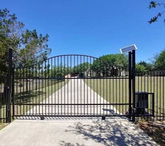 Black metal driveway gate with solar panel and motor on a paved driveway, sunny day.