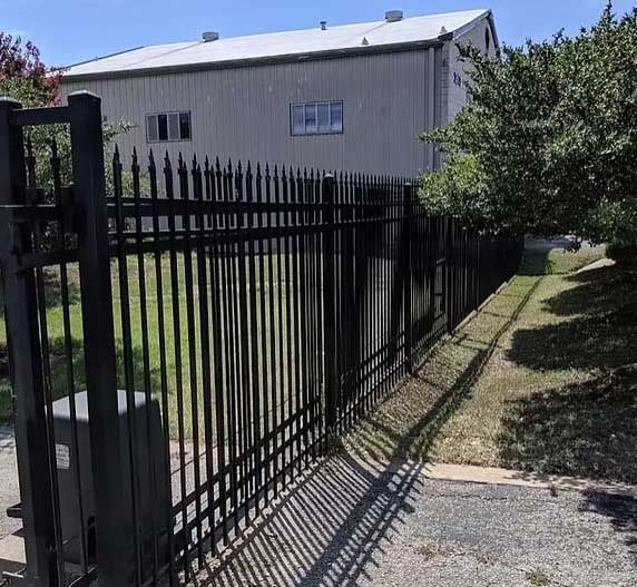 Black metal fence surrounds a light gray building with two windows. Green grass and trees are visible.