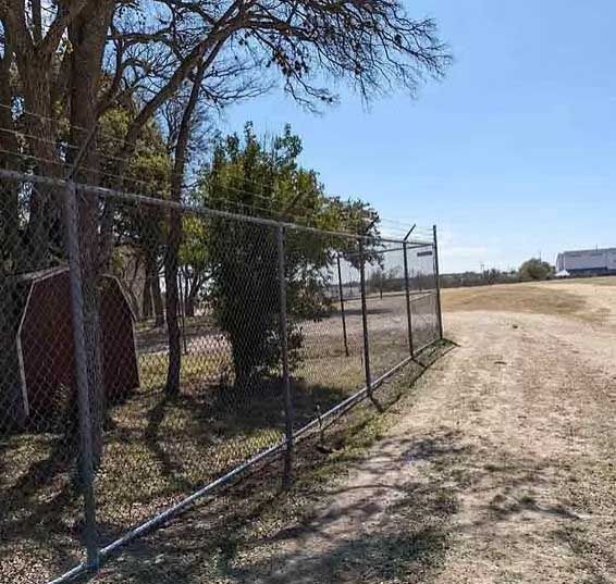 Chain-link fence with barbed wire surrounds a grassy field. A small shed and trees are on the left.