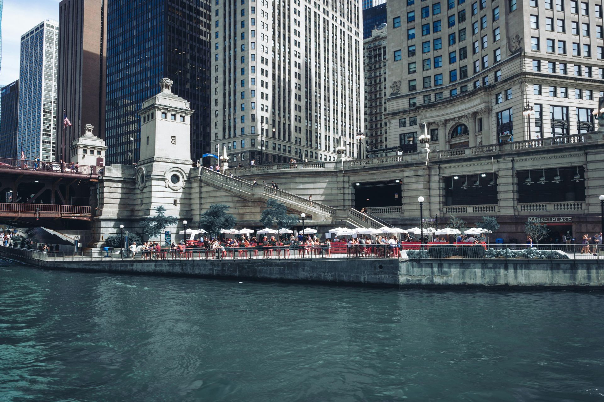 Chicago Riverwalk with tall buildings and bridge towers. Water reflects the city.